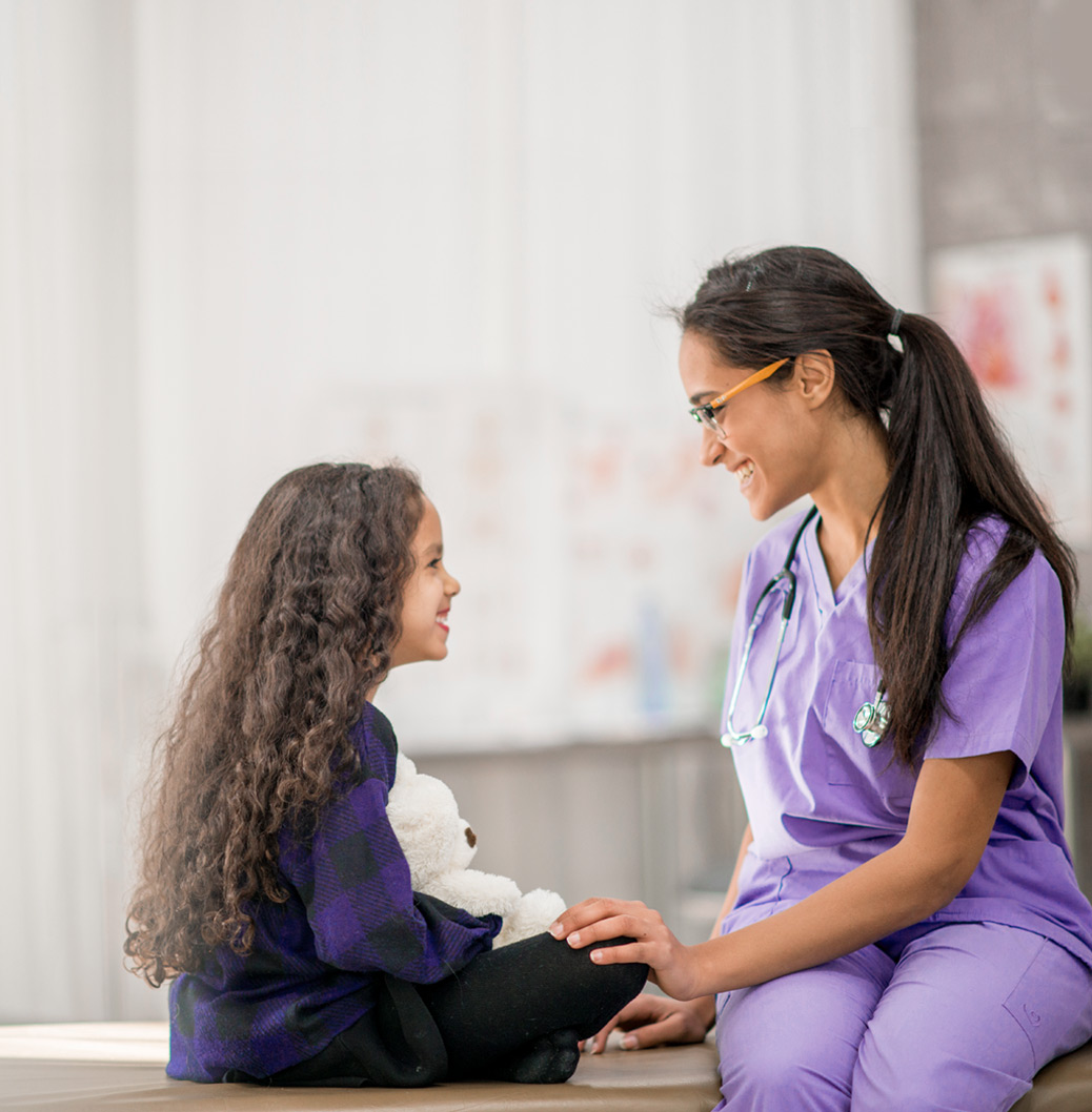 Medic pediatru în uniformă mov zâmbește unei fetițe cu păr creț care ține un ursuleț de pluș